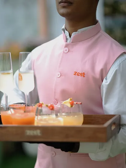 A server in a sharp pink "Zest" uniform holds a tray of welcome drinks.