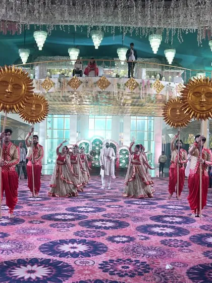 The stage is set for a royal groom entry. Dancers holding large sun props create a grand formation on a patterned carpet, awaiting the groom's arrival at this opulent wedding venue.