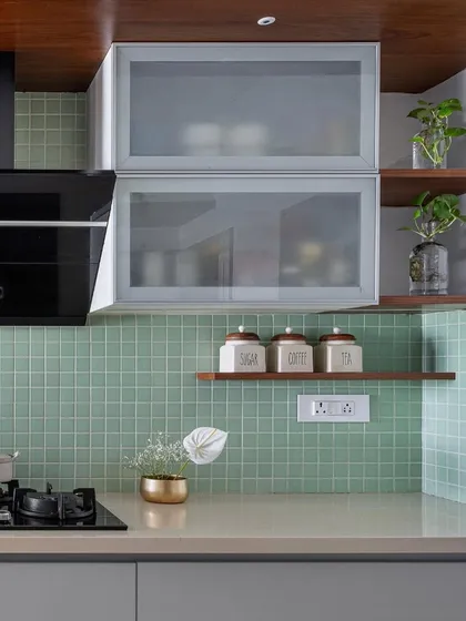 A close-up of the 'Gulmohar' kitchen palette, featuring mint green tiles, grey cabinets, and frosted glass overhead units. The wooden ceiling panel adds a layer of warmth.