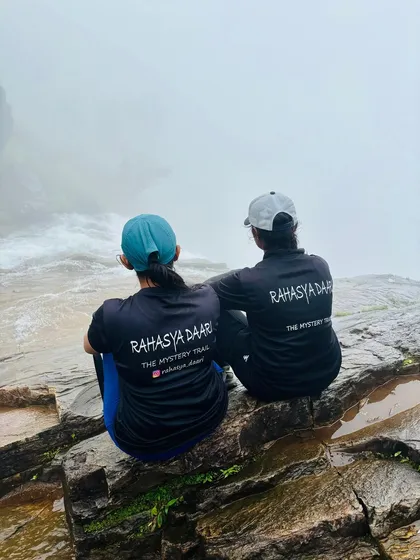 Two of our trekkers in Rahasya Daari t-shirts sitting peacefully by the edge of a misty waterfall, a moment of quiet reflection.