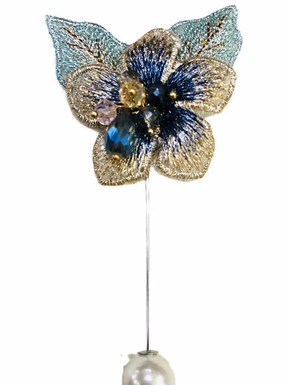 This lapel pin showcases an intricately embroidered flower in shades of gold and blue, accented with crystals and a pearl drop. It is a small piece of art for your attire.