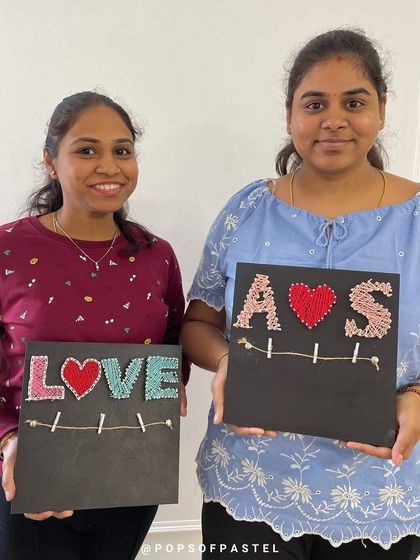 Two friends display their finished string art, one with the word "LOVE" and the other with personalized initials.