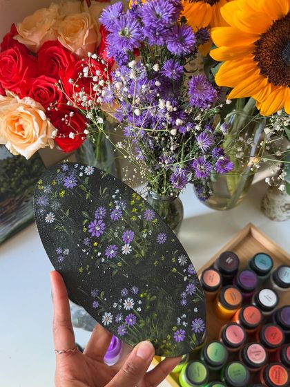 Here, the finished oval painting is shown with the fresh purple flowers that inspired it, along with my collection of paints. It connects the inspiration, the tools, and the final art.