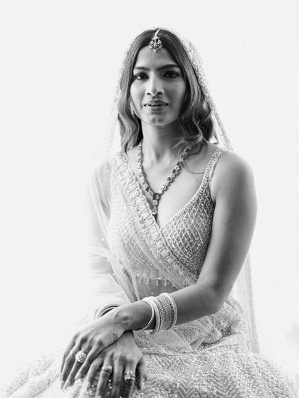 A classic black and white bridal portrait. The high-key lighting creates a soft, ethereal glow, highlighting the bride's features and her beautiful white lehenga.