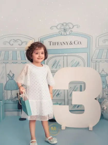 A stylish toddler celebrating her third birthday with a "Tiffany & Co." theme, holding a shopping bag and standing next to a large number "3".