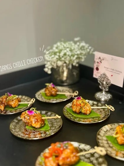 Jiang's Chilli Chicken bites on skewers, placed on banana leaves atop golden plates, blending traditional and contemporary elements.
