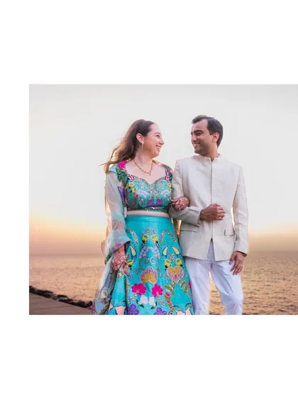 A beautiful portrait of the couple by the sea at sunset. The warm colors of the sky complement her vibrant outfit, creating a stunning visual.