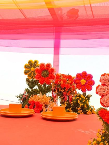 A close-up of a colorful floral centerpiece shaped like a flower. The use of bright red, orange, and yellow flowers on the tabletop adds a playful and artistic touch to the haldi decor.