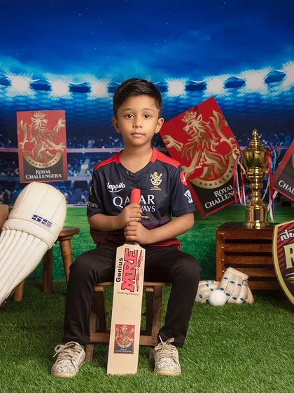 A true RCB fan! This young cricketer is ready for the match, surrounded by team flags and holding a bat with determination.
