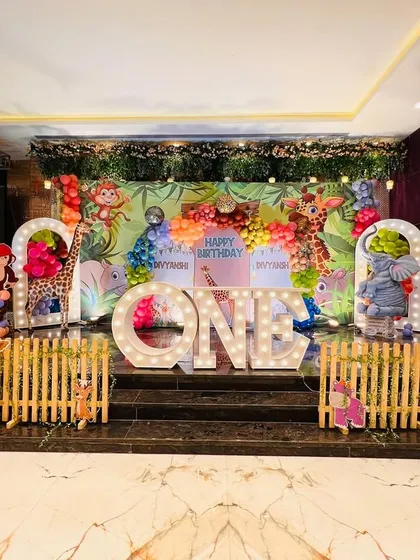 A complete jungle safari stage setup inside a banquet hall, featuring a wooden fence, animal cutouts, and "ONE" marquee letters.