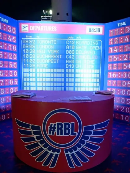 A travel-themed installation featuring a large airport-style departure board and a branded check-in counter, designed for a corporate event.