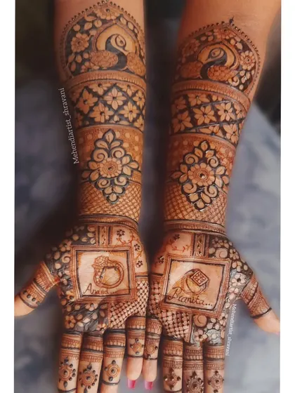 A clear, still image of the engagement ring mehendi. The names are clearly visible, and the design is complemented by traditional peacock motifs on the forearms.