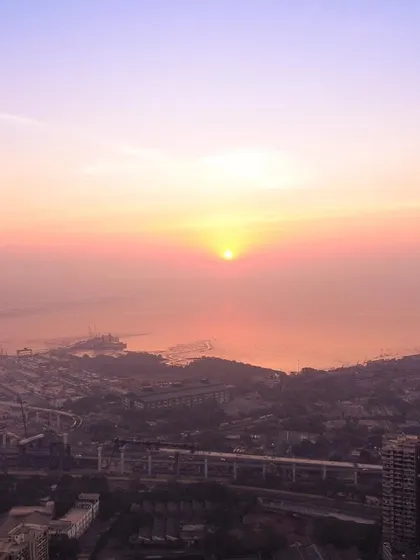 A hazy sunrise over Mumbai, with the sun appearing as a bright orb above the Bandra-Worli Sea Link and the coastline.