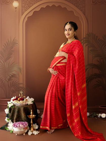 A full-length portrait of a mom-to-be in a beautiful red saree, standing next to a table with traditional offerings, looking directly at the camera with a gentle smile.