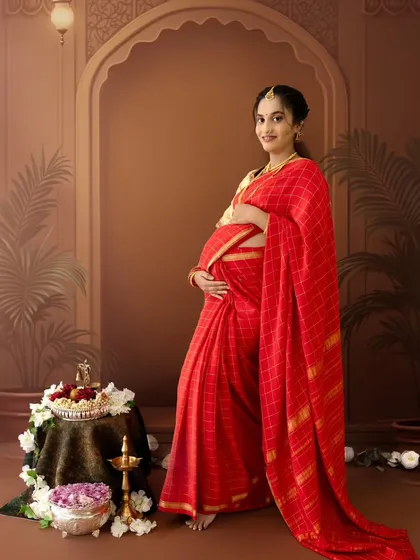 A full-length portrait of a mom-to-be in a beautiful red saree, standing next to a table with traditional offerings, looking directly at the camera with a gentle smile.