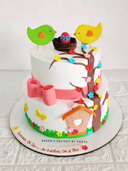 A cheerful two-tier baby shower cake with a bird theme. It features two lovebirds, a nest with an egg, a birdhouse, and a bright pink bow, perfect for welcoming a new baby.