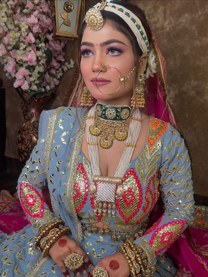 A detailed portrait showcasing the bridal makeup and jewellery. The green choker stands out beautifully against the blue outfit, and the pearl-lined matha patti frames the face perfectly.