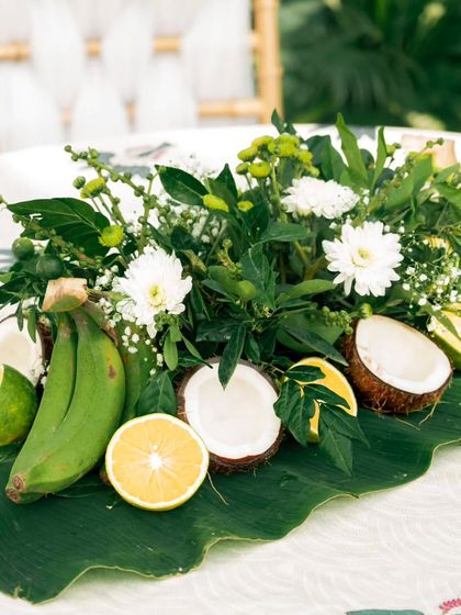 A detailed look at a natural table centerpiece, featuring fresh coconuts, bananas, oranges, and white flowers on a large leaf. This type of decor is perfect for traditional or tropical-themed events.