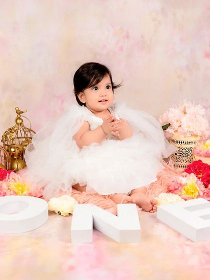 A baby girl in a fluffy white dress celebrates her first birthday with "ONE" blocks and a beautiful pink floral backdrop.