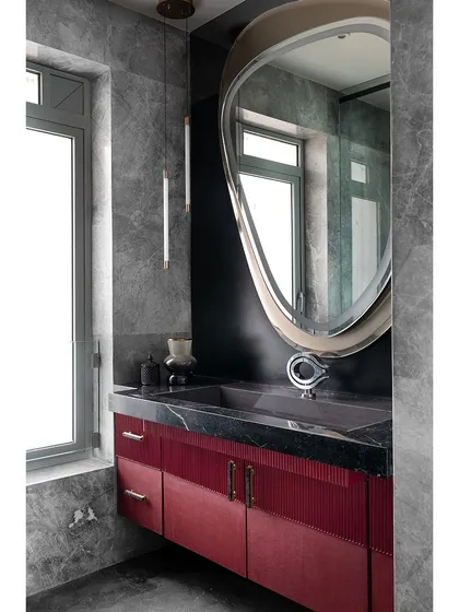 In this master bathroom, a rich red vanity with fluted detailing adds a pop of color against the grey marble walls. The organically shaped mirror and sleek pendant light enhance the modern, luxurious feel of the space.