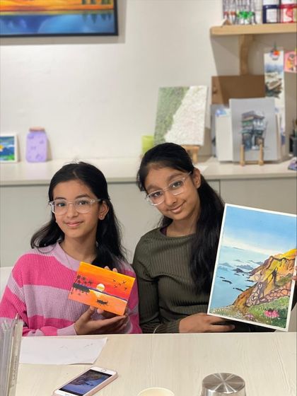 Two sisters displaying their different creations from a single session. One chose to paint a sunset on a small box, while the other created a detailed landscape on paper.