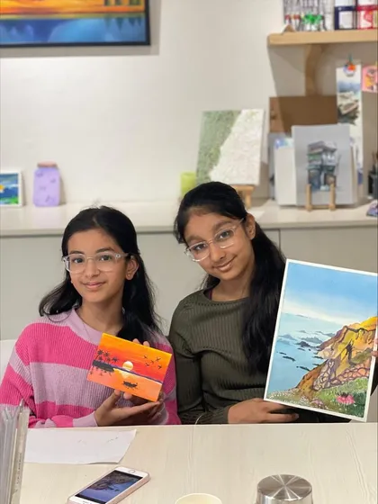 Two sisters displaying their different creations from a single session. One chose to paint a sunset on a small box, while the other created a detailed landscape on paper.