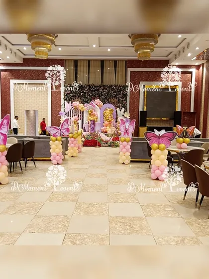 A full view of the banquet hall decorated for a butterfly-themed party. The walkway is lined with balloon pillars topped with large butterfly wings, leading guests towards the main stage and creating an immersive experience.