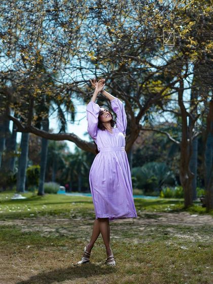 A model in a lavender dress reaches towards the sky, creating a sense of aspiration and movement. The wide-angle shot emphasizes the open, natural space.