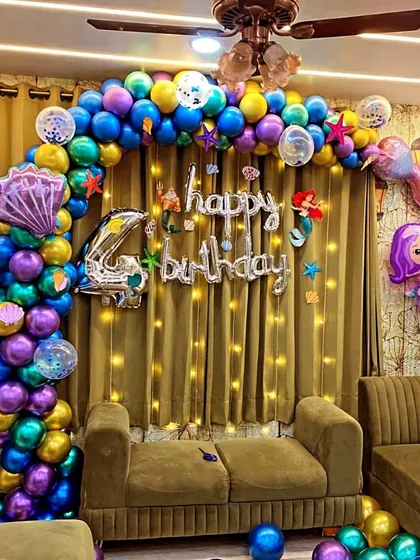 An elaborate mermaid-themed birthday setup in a living room. A full balloon arch with under-the-sea colors frames the sofa, complemented by fairy lights and character foil balloons.