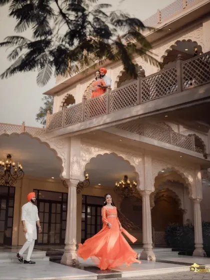 A wide-angle shot that captures the scale of the heritage location. The couple on the balcony and the bride's flowing orange dress create a dynamic and visually stunning composition.