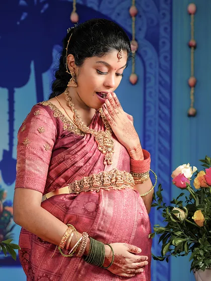 A candid shot capturing the wonder of pregnancy. The mom-to-be, adorned in a pink silk saree and gold Oddiyanam, shares a sweet, surprised expression during her baby shower event.