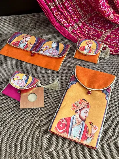 A set of Raja-Rani themed wedding accessories, including shagun envelopes and coin pouches. The vibrant orange and pink colors are perfect for a festive celebration.