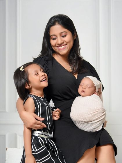 A mother holds both her children, capturing a moment of pure happiness. The older daughter's joyful laugh beside her sleeping newborn sibling perfectly illustrates the love in a growing family.