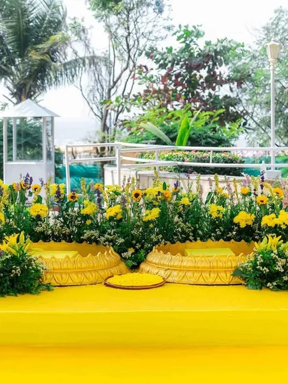 A simple and elegant Haldi seating area by the sea. The stage is covered in bright yellow, with two traditional "bajots" for the ceremony, surrounded by beautiful sunflower arrangements.