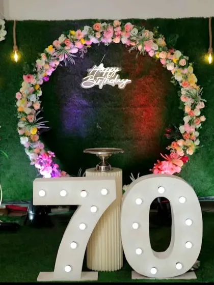 A stylish 70th birthday celebration with a circular floral arch against a lush green backdrop. The setup is highlighted by large, illuminated marquee numbers and elegant gold stands, making it a perfect photo opportunity for a major milestone.