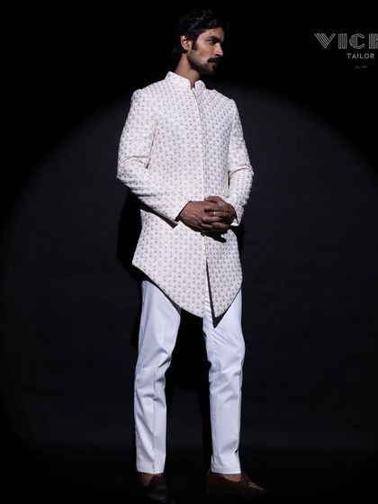 Modern architecture in tailoring. This white sherwani features a striking asymmetric hemline and all-over geometric embroidery, crafted for the groom who appreciates contemporary design.