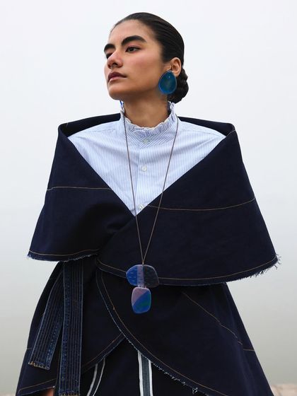 A close-up on the styling, showing how a simple white shirt is layered under a draped denim piece. The sculptural jewelry adds a touch of modern artistry.