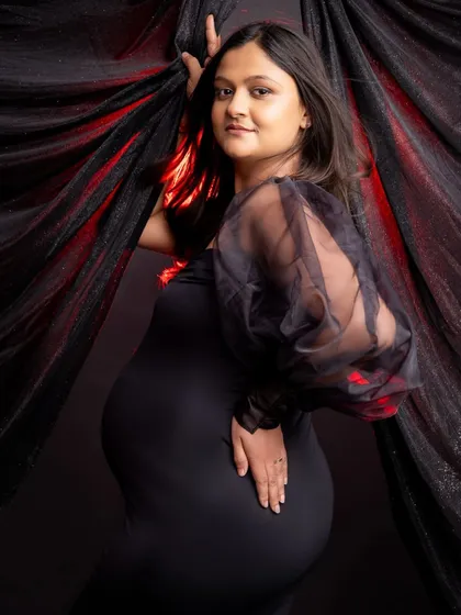 A classic maternity pose against a dark, textured backdrop. The expecting mother, in a fitted black dress with sheer sleeves, gently holds the fabric, looking confidently at the camera.