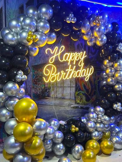 A cool and stylish balloon ring decoration for a birthday, using black, silver, and gold balloons. Set against a graffiti wall, this backdrop with a 'Happy Birthday' neon sign is perfect for a teen or adult party.