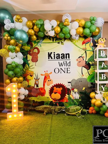 A full view of Kiaan's "Wild One" jungle birthday setup. The backdrop is filled with cartoon safari animals, framed by a lush green and gold balloon garland, and accented with a light-up number '1' and 'BABY' blocks.
