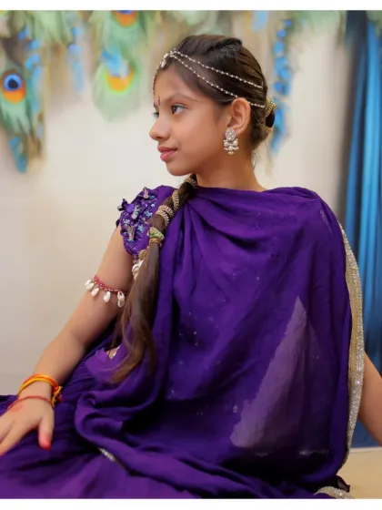 A thoughtful profile shot of a girl dressed as Radha, highlighting the traditional jewelry and braided hairstyle against a peacock feather backdrop.