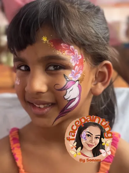 A smiling girl proudly showing off her unicorn and floral face paint. The red glitter adds a beautiful pop of color.