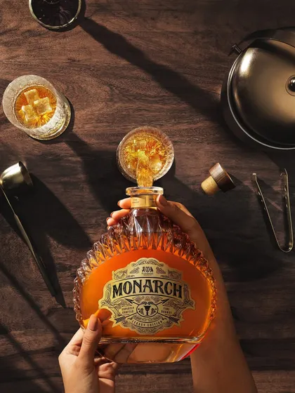 A wider angle of the top-down pour for Monarch. This view includes more of the surrounding scene, with the bottle, glasses, and bar tools creating a complete story of someone preparing a drink.
