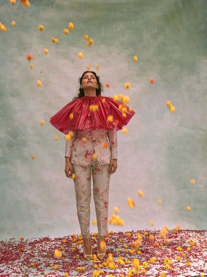 A surreal editorial image featuring a floral suit and a dramatic red pleated collar. The styling creates a unique, artistic silhouette, enhanced by the shower of falling flowers against a painterly backdrop.