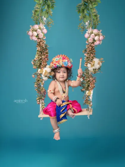 A serene portrait of a baby as Krishna on a floral swing, holding a flute. The solid blue background creates a calm and dreamy atmosphere for this divine-themed shot.