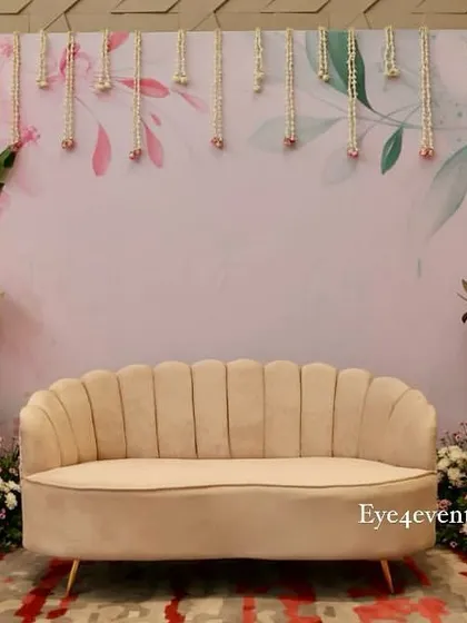 This beautiful backdrop was designed for an engagement party, featuring a soft, floral-printed background with hanging floral strings. The setup is framed by tropical leaves and flower arrangements, with a plush beige sofa for photos.