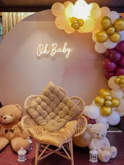 A cozy and elegant photo corner for a baby shower. This setup features a comfortable peacock chair for the mom-to-be, flanked by teddy bears and a beautiful balloon arrangement with a simple "Oh Baby" neon sign.
