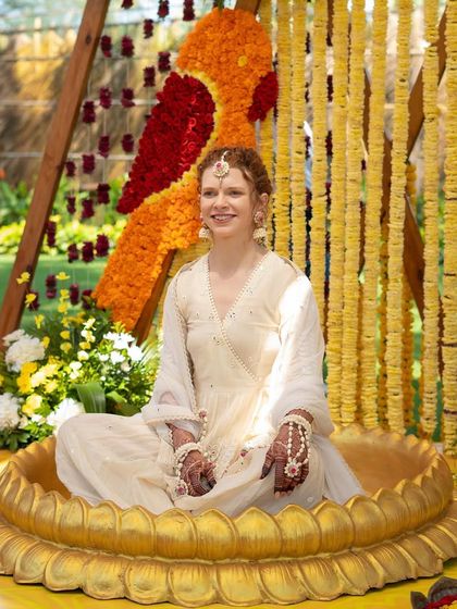 The bride sits in a lotus-shaped vessel for the Haldi ceremony. Her custom-made ivory sharara set is accessorized with delicate floral jewelry, adding a soft touch of pink.