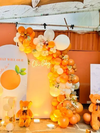 A bright and cheerful 'Little Cutie' baby shower with an orange and teddy bear theme. The main backdrop features a large balloon arch and a neon 'Oh Baby' sign.
