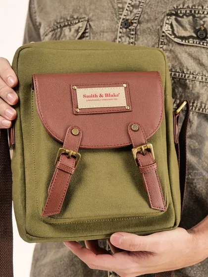 A man holding an olive green and brown canvas messenger bag. This shot focuses on the bag's details, like the buckles and brand plaque.